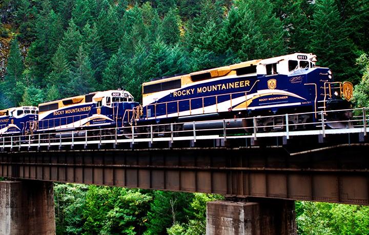 Blue train traveling across a bridge with woods behind, text on train says "Rocky Mountaineer." says, "