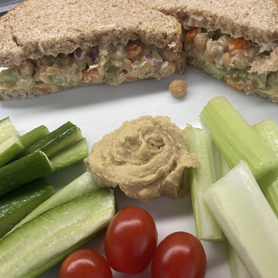 A white plate containing a chickpea salad sandwich, raw vegetables and hummus.