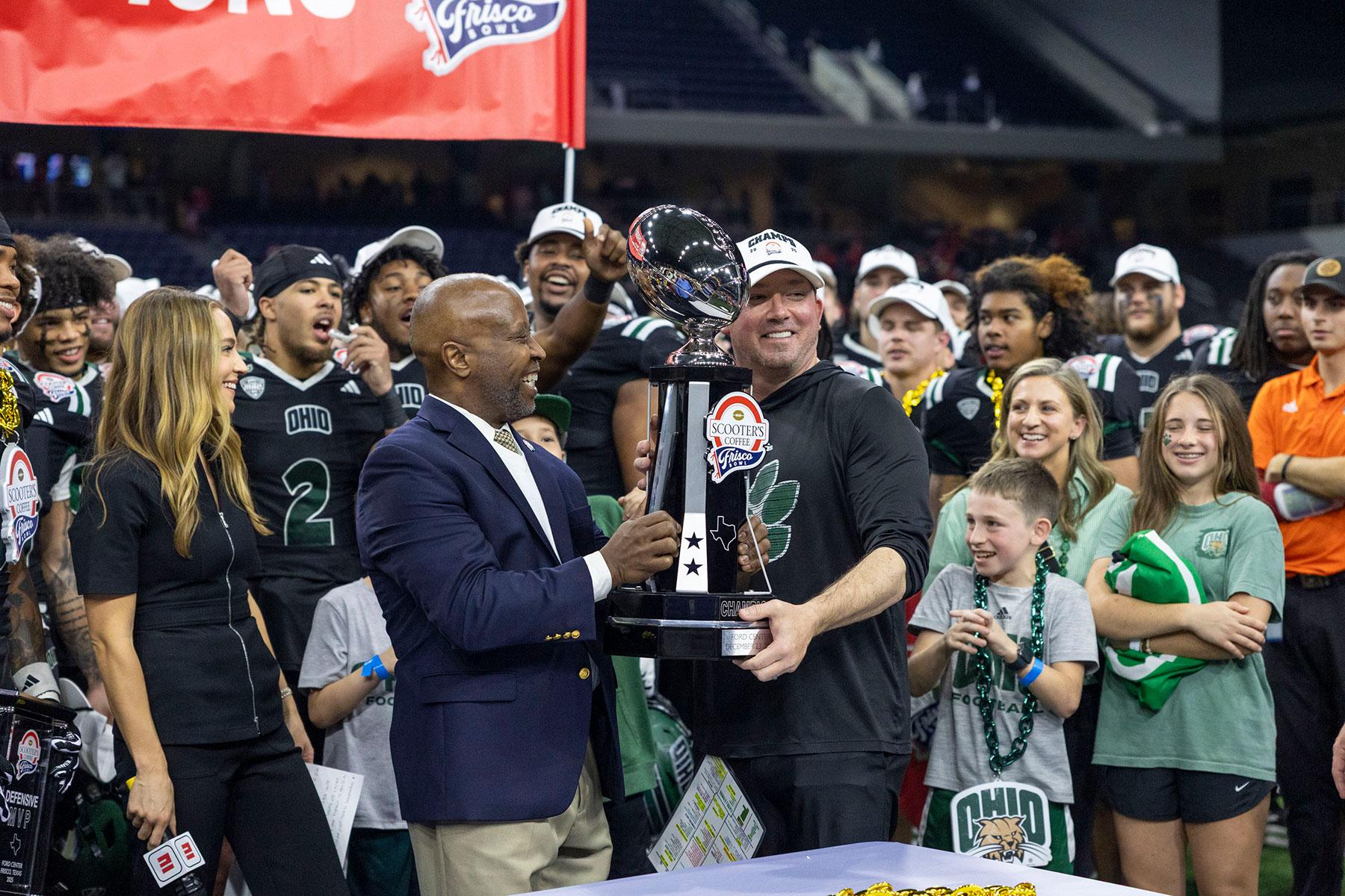 OHIO football coach holds the trophy for the 2025 Frisco Bowl after the game