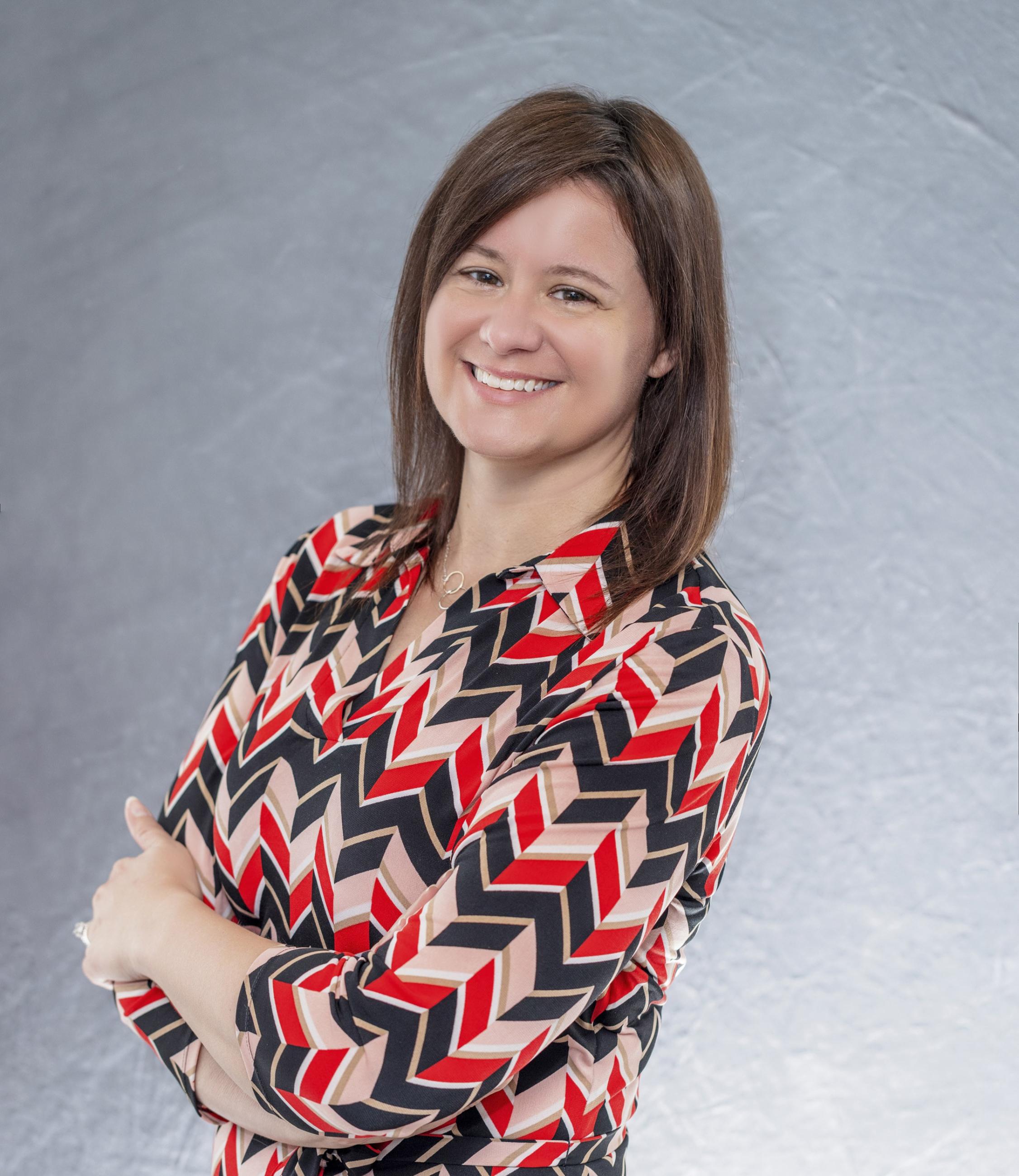 a white woman smiling facing the screen with shoulder length brown hair wearing a black and red chevron pattern top