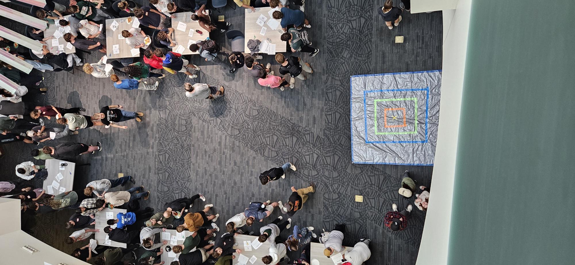 Students standing in an oval around a mat where an egg-drop competition is taking place