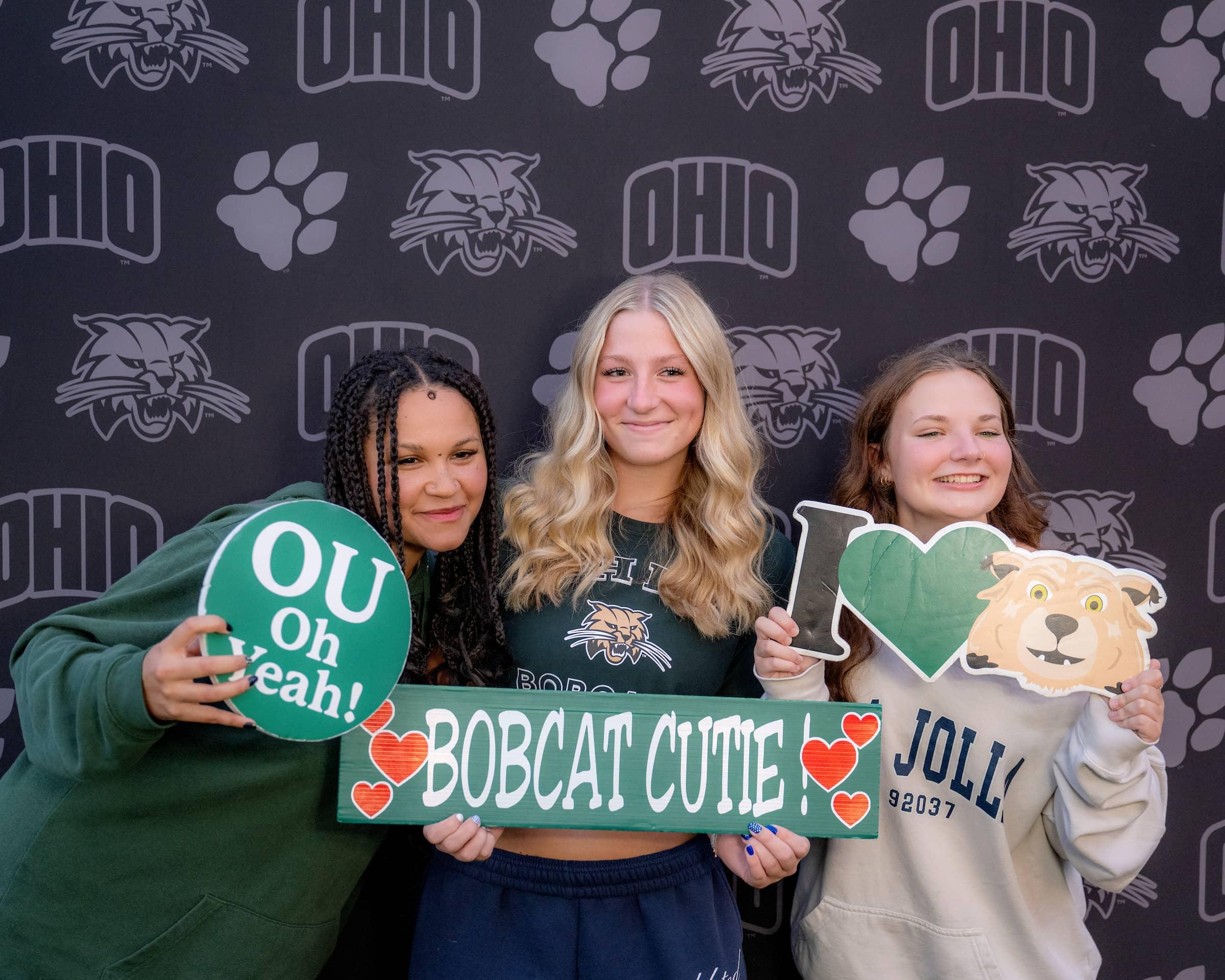 3 students pose in a photo booth holding sign props "I love bobcats" "OU Oh Yeah" "Bobcat Cutie"