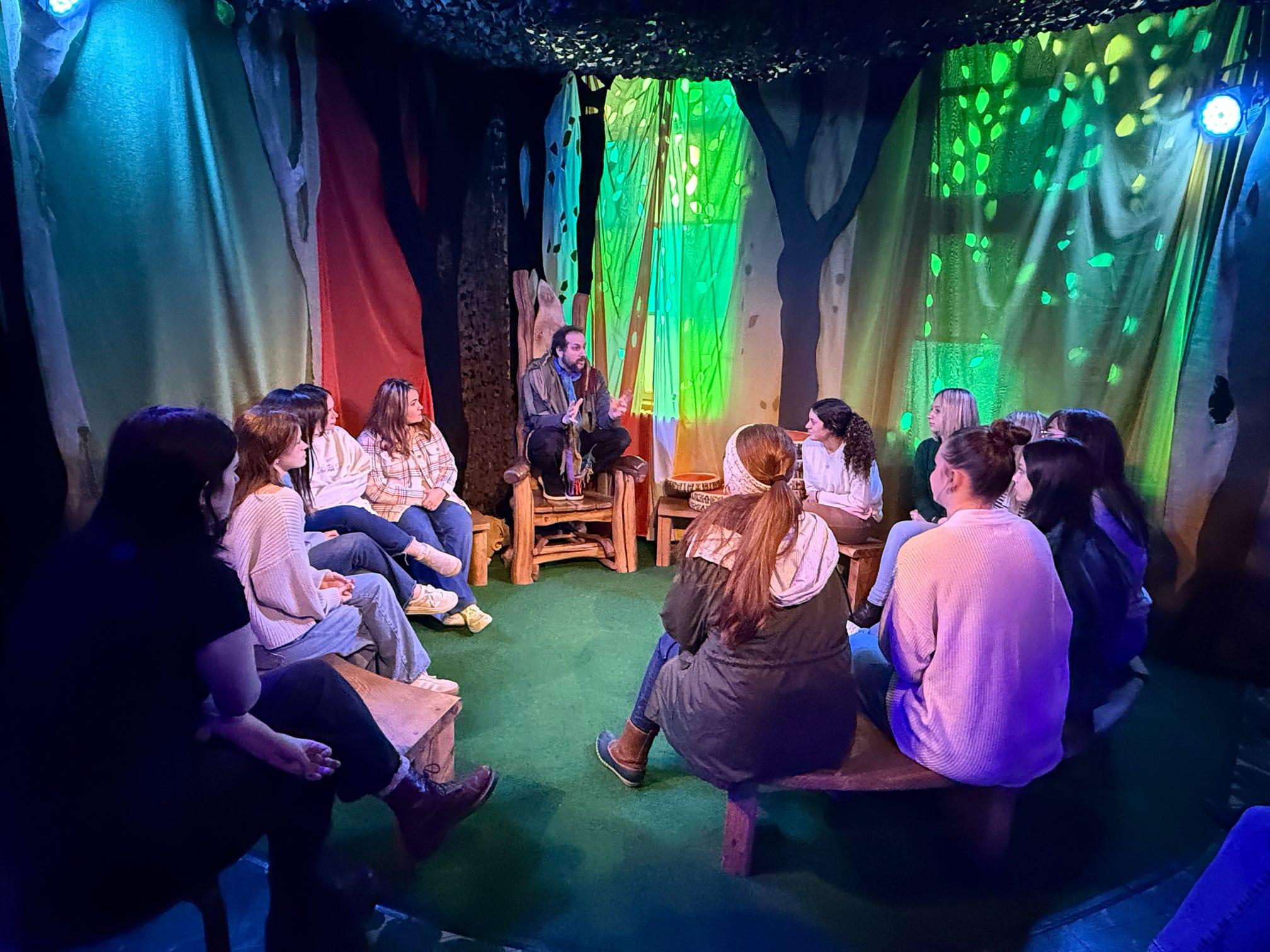 A group of Ohio University students sit in a circle in a blue and green lit room with fairy lights and colorful curtains at a children's literature museum