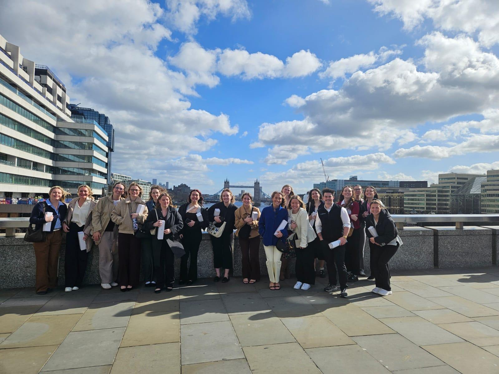 A group of Ohio University Students and Faculty pose on a bridge with the London Tower Bridge in the background with a blue sky and clouds.