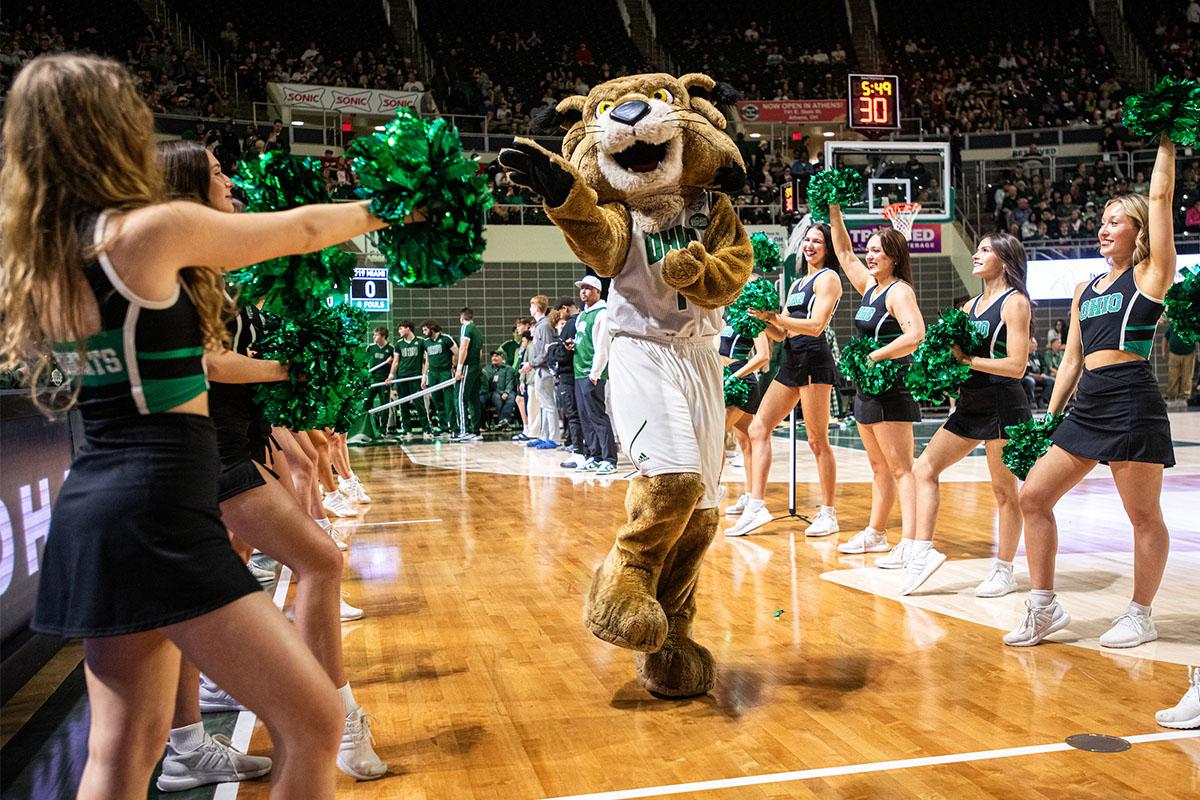 OHIO's mascot, Rufus the Bobcat, skips while reaching towards a cheerleader.