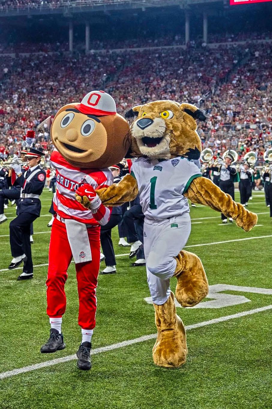 Ohio University's mascot, Rufus the Bobcat, and The Ohio State University's mascot, Brutus Buckeye, hold hands and skip together across the football field.