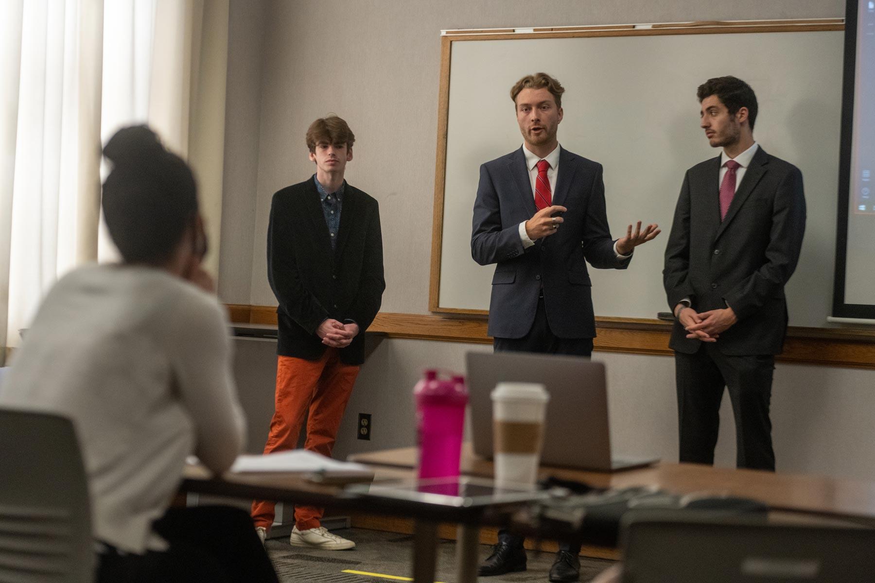 three students wearing suits give a presentation in the front of a classroom