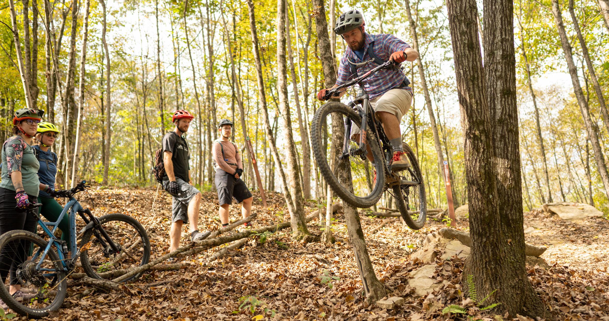 man jumps with mountain bike in the air while companions watch in beautiful forest