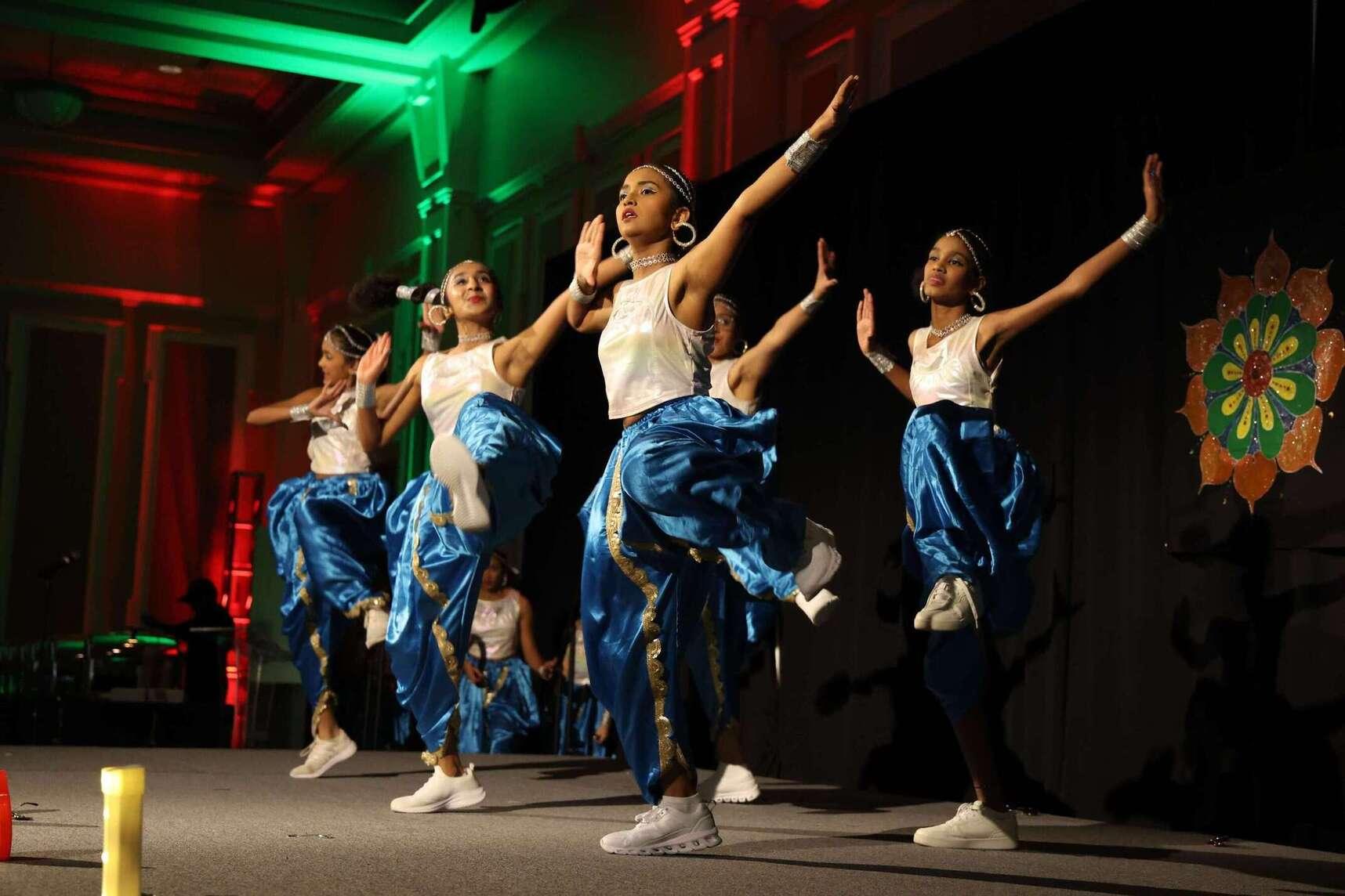 Group of Sai Arts students performing wearing blue satin pants with one long silver stripe down each leg, and white satin tank tops. All of them have their arms in the air kicking one leg out in motion