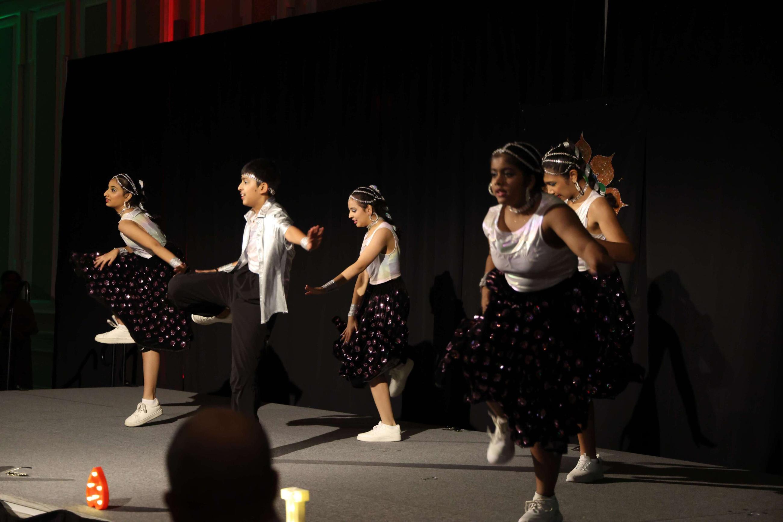 Five members of the Sai arts group wearing black skirts and pants and white shirts kick their legs up as they perform a dance. Each are wearing silver beads on their heads