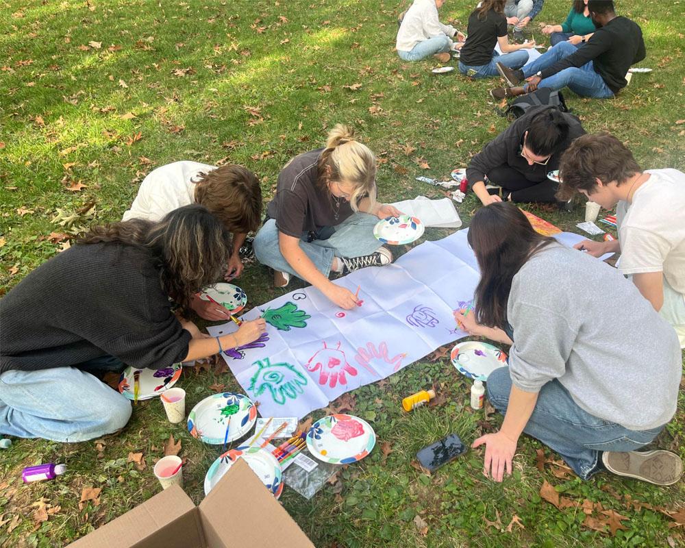 group of students sit on the grass on college green painting hands on a big banner