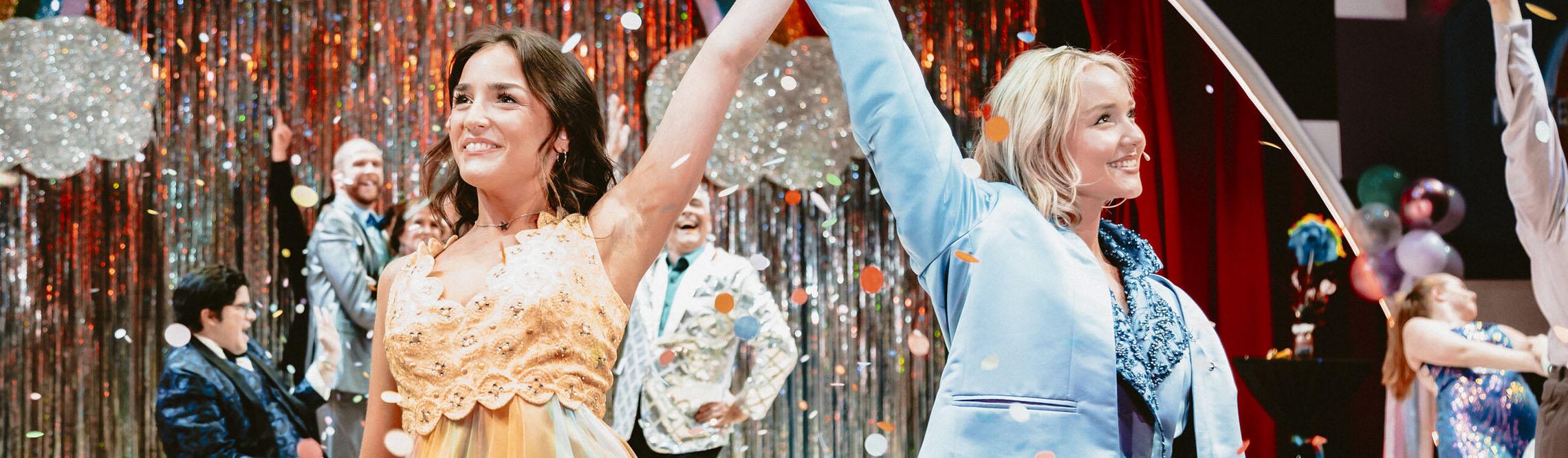 two female leads raise their held hands in front of a theater stage