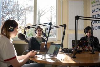 three students sit in front of microphones in the podcast studio