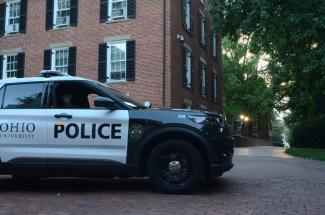 Police vehicle marked "OHIO UNIVERSITY POLICE" parked on a brick pathway beside a red-brick campus building with white-trimmed windows and black shutters.