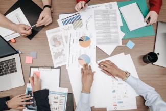 Hands of four people around a table that has printed pages of charts, data, etc.