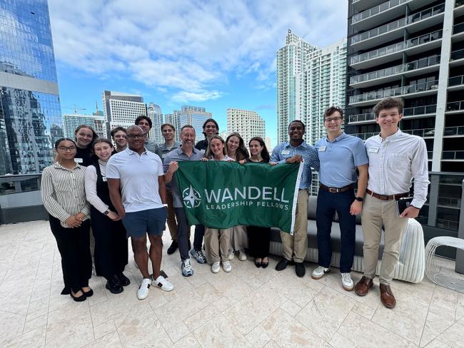 Several students in the Wandell Fellows pose on top of a skyscraper, holding a flag for the Wandell Fellows group