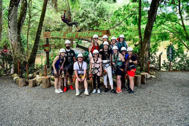 Group standing in front of a sign "Pura Vida Paradise" in zip lining gear