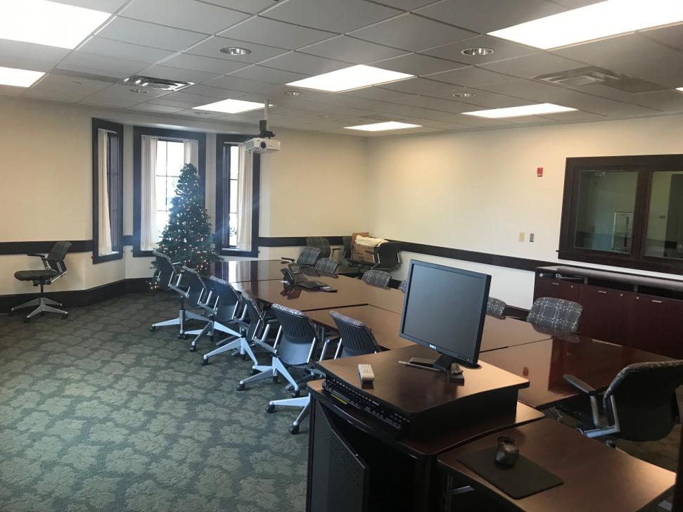 Conference room with several wood tables pushed together to form a larger one, surrounded by about 15 wheeled chairs. A computer and A/V cabinet nearby. A Christmas tree near the end of the table by the windows.