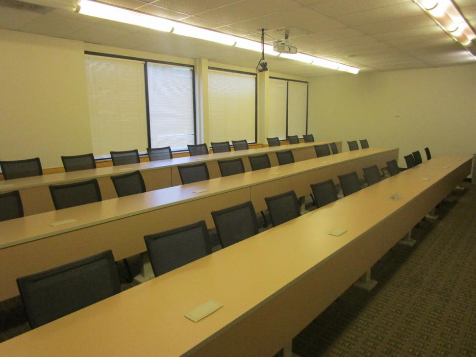 Lecture hall with three long desks extending across the room, about 30 chairs, projector suspended from ceiling