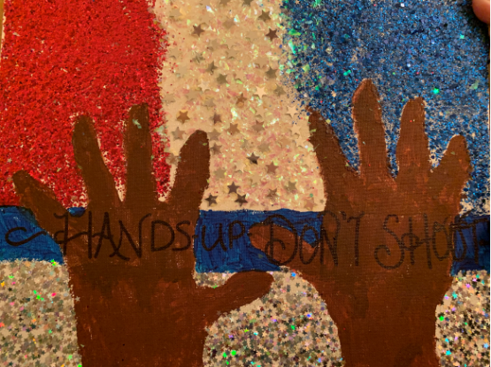 In the picture above a woman's hands are held up with the phrase “Hands up, don’t shoot.” The background is red, white, and blue to symbolize the lights of a police car.