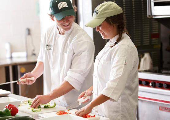 two students wearing white coats preparing food 