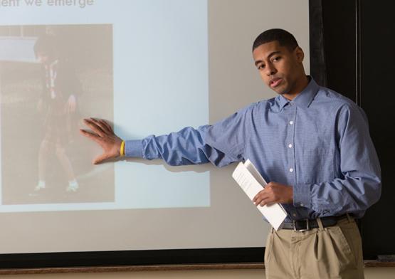 A man stands in front of a projector screen while motioning to the images being displayed on the screen