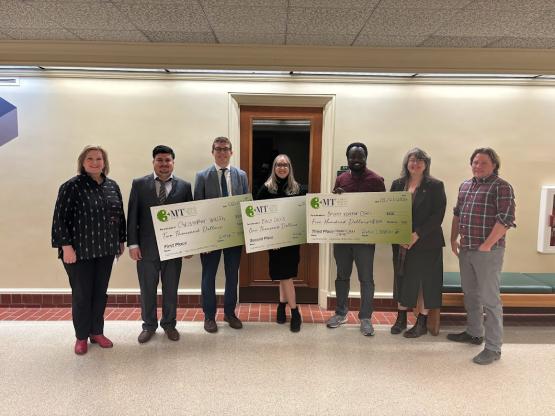 A group of people standing in a hallway, three holding large checks