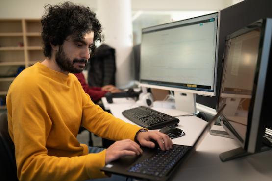 A man with curly dark hair and a beard is working at a desk in an office setting. He is wearing a yellow sweater and typing on a laptop.