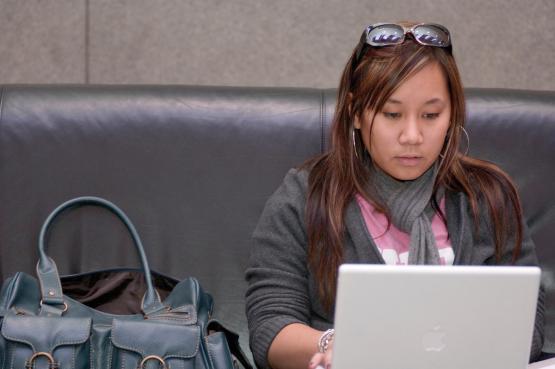 Woman working on a laptop. She is wearing a grey jacket, a grey scarf, and sunglasses on her head. 