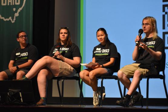 Four students in OHIO BSO T-shirts sit on a stage. One is holding a microphone and speaking while the other three listen. 