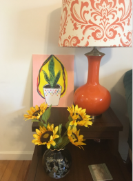 A painting of a plant is on a table with a lamp and vase filled with sunflowers.