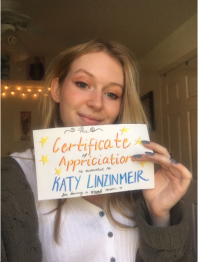 Taylor holding a piece of paper that reads’ “This Certificate of Appriciation is awarded to Katy Linzinmeir for being a great mom” in orange and blue marker.  