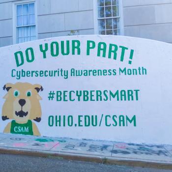 A white wall painted with green text saying "Do Your Part! Cybersecurity Awareness Month #CyberSmart" and a Rufus Bobcat with a CSAM shirt.