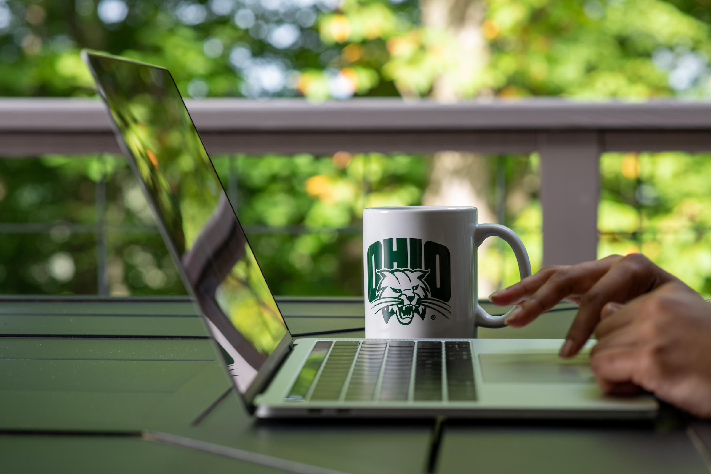 Laptop viewing away to the side, with hands typing away on the keyboard. A white mug with a green Ohio University symbol is seen behind the computer. 