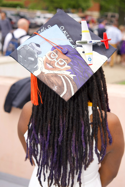 A young woman faces away from the camera, with her cap displayed with including a drawing of her as a pilot, a toy plane attached, with the words "Laurél D Class of '19" on it