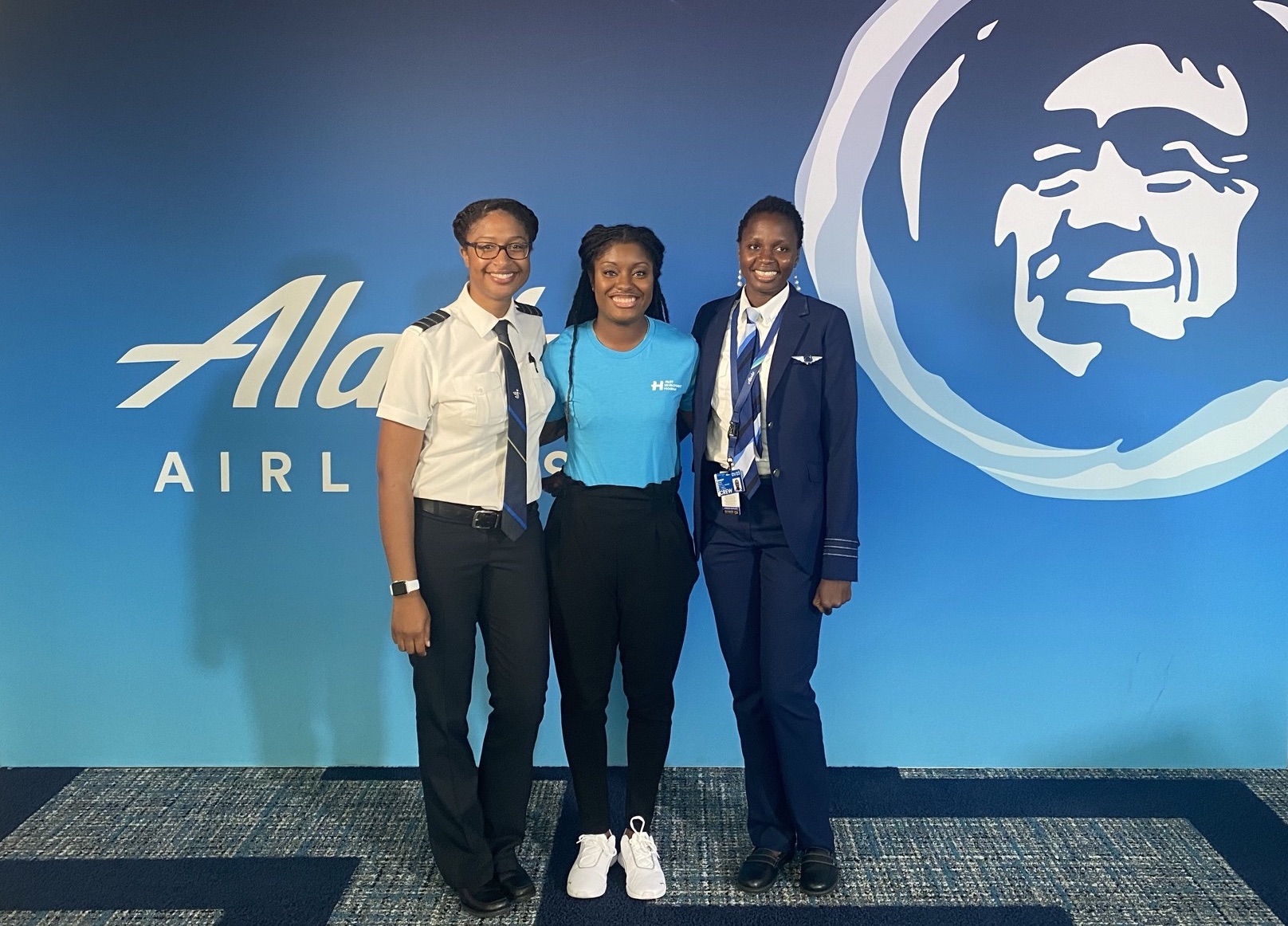 Three women stand together smiling in front of an blue background with Alaska Airlines on it