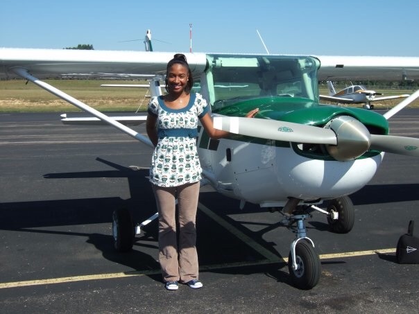 A young woman stands in front of a small green and white plane.
