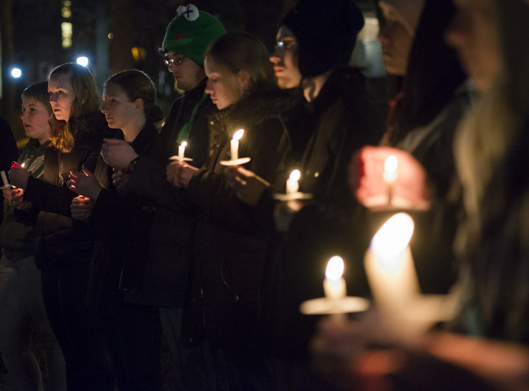 A group of men and women, huddled together, stand at night holding candles as they look off camera towards the right.
