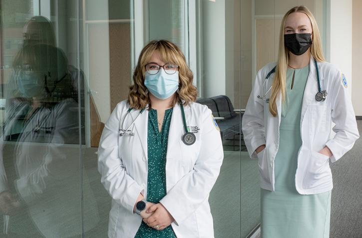 Two women wearing lab coats and masks look at the camera