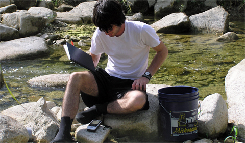 A young Aiden Shearer sits on a large rock on the edge of a creek with a bucket on the ground and a notebook in his hand