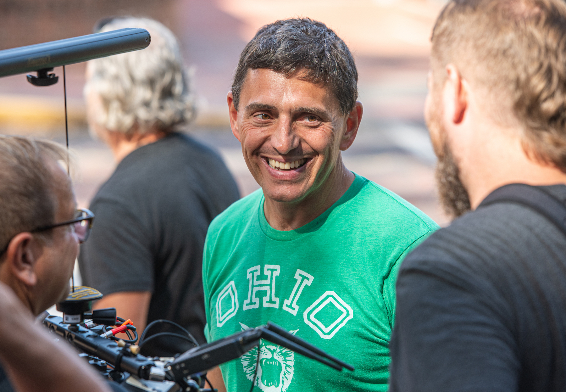 A man wearing a green Ohio University shirt smiles with people huddled around him