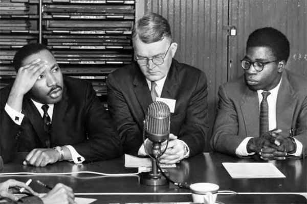 This image appears in the Ohio Alumnus, January 1960, captioned: "Each day while the Conference was in session, a press conference was held each morning with newsmen and photographers from newspapers, radio and television attending. Above Martin Luther King answers a question for the newsmen. Seated beside Rev. King is Dr. Winburn Thomas, leader of the forum on racial tensions. Mr. 'Bola Ige, co-secretary of the conference, which is sponsored by the National Student Christian Federation, in on the right." (