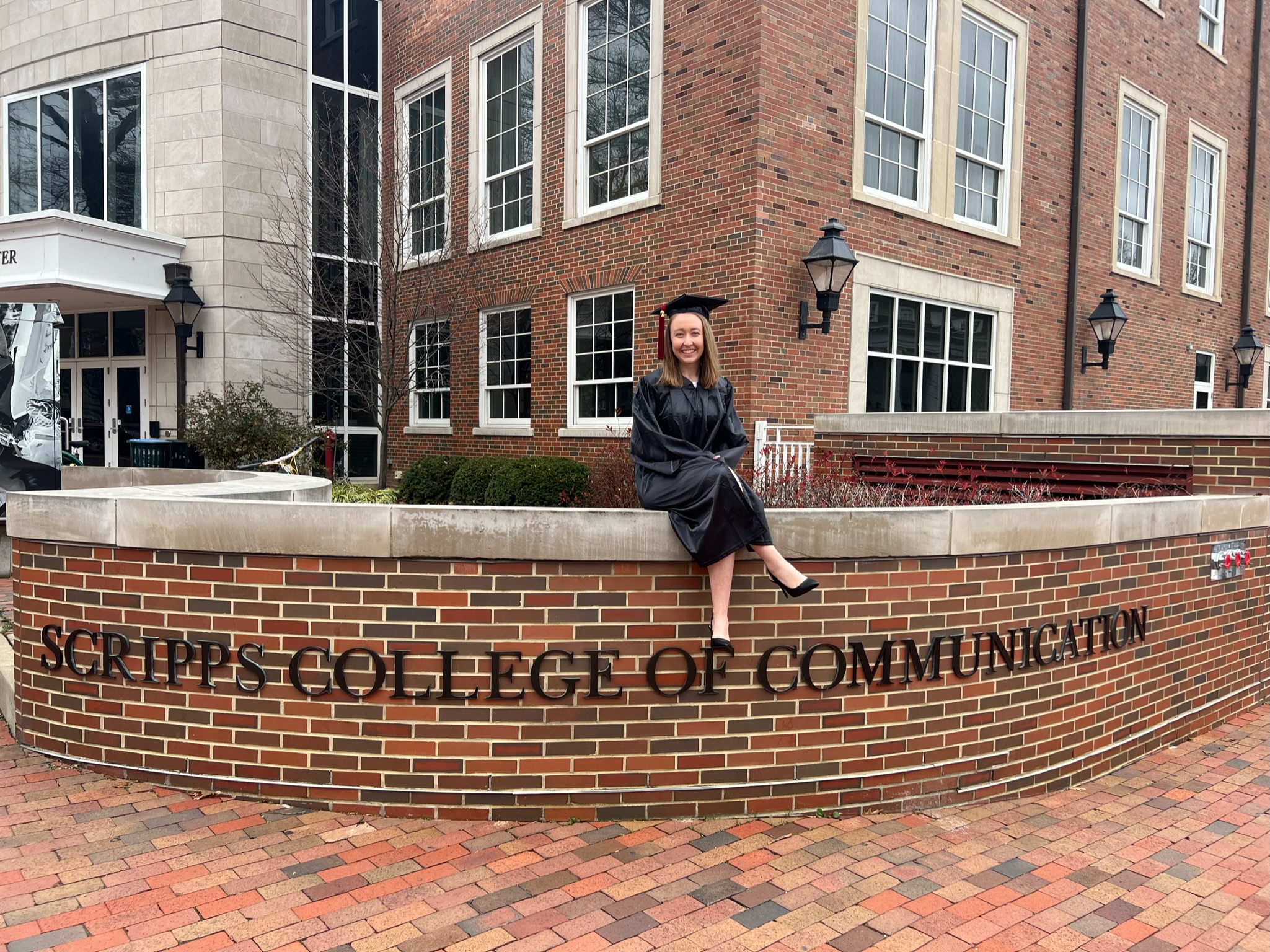 Kirsten Thomas sits on a side wall that reads "Scripps College of Communication" on the Ohio University Athens Campus