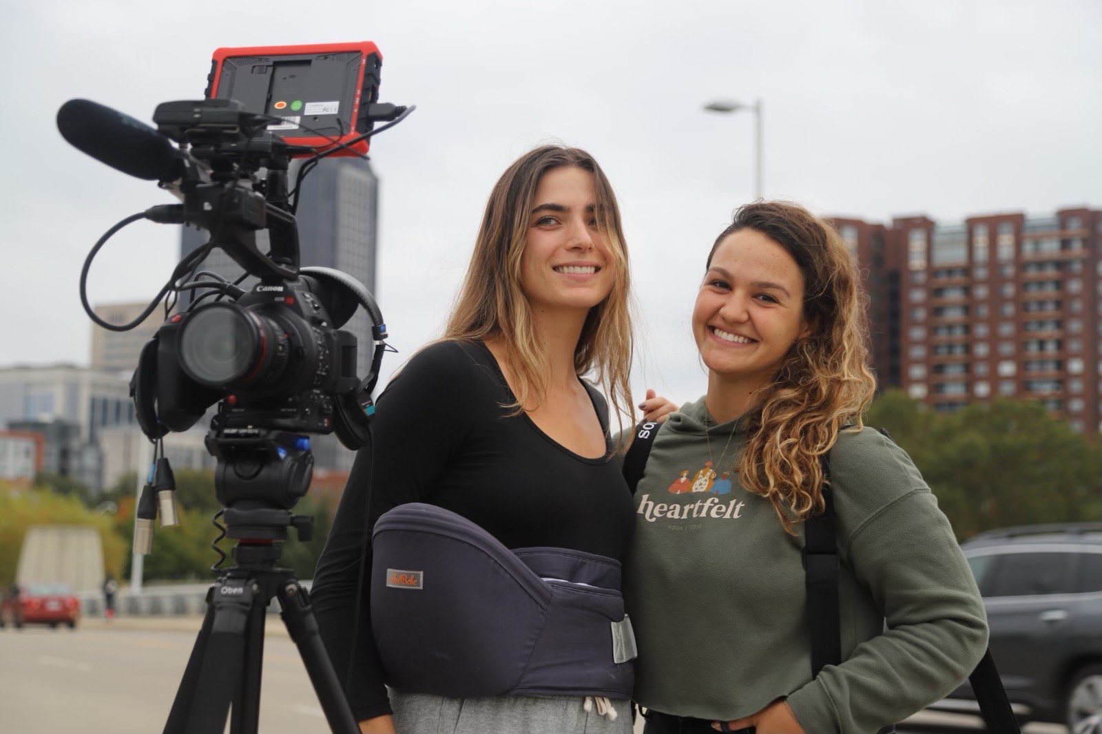 Two women pose for a picture behind a camera with city buildings in the background.
