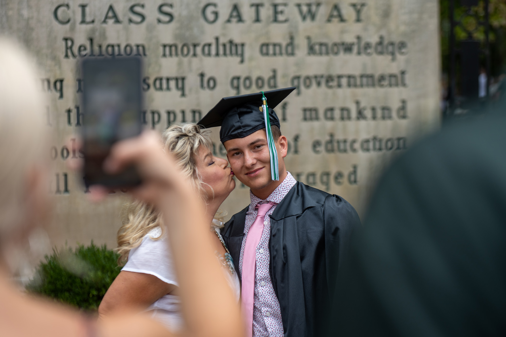 A graduating student poses for a photo in front of the Class Gateway while his mother kisses his cheek