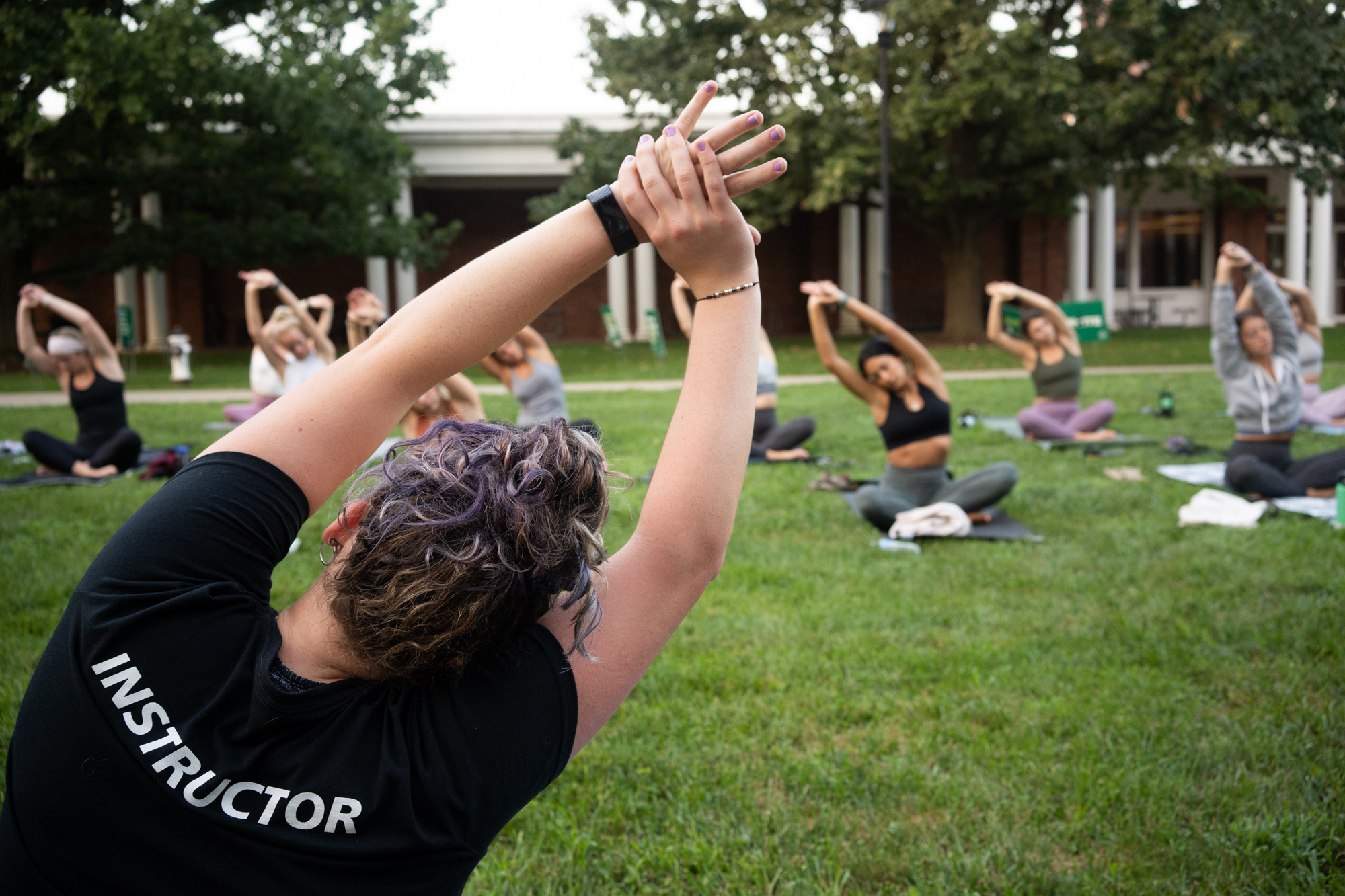 The back of a yoga instructor is visible leading seated stretches on West Green