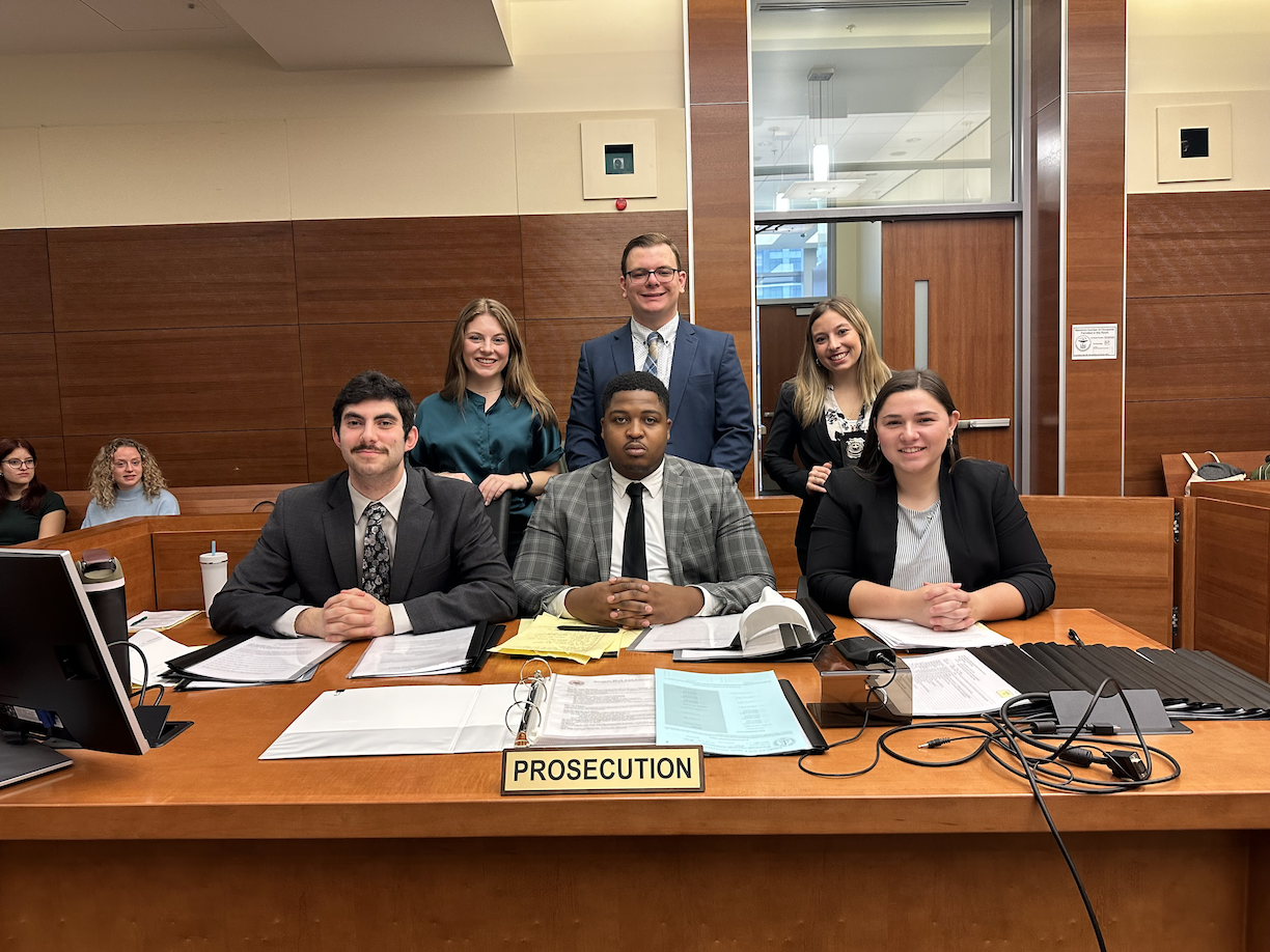 A group from Ohio University's Mock Trial Team poses in a courtroom, seated at a table behind a placard that says "Prosecution"