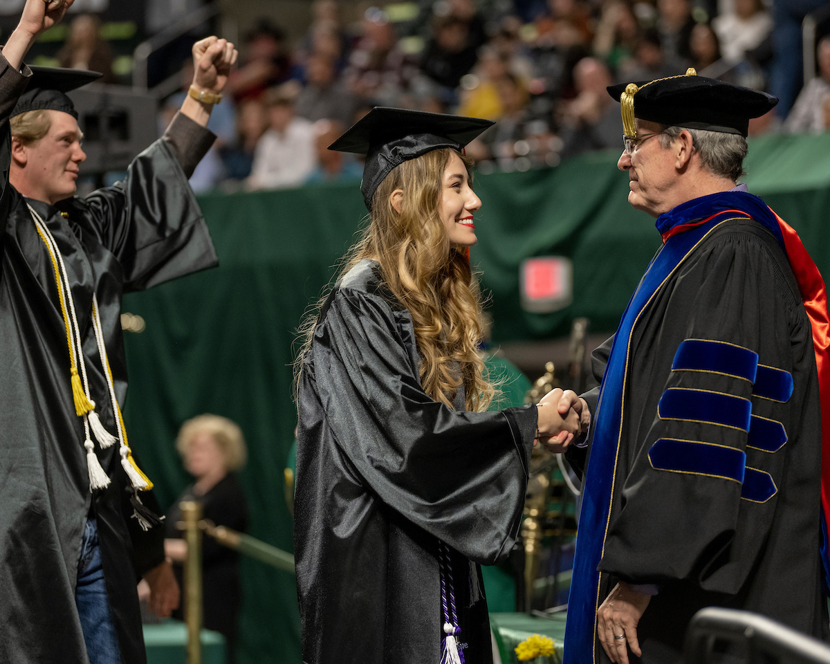 Female graduate is congratulated onstage while faculty behind her raises hands in celebration