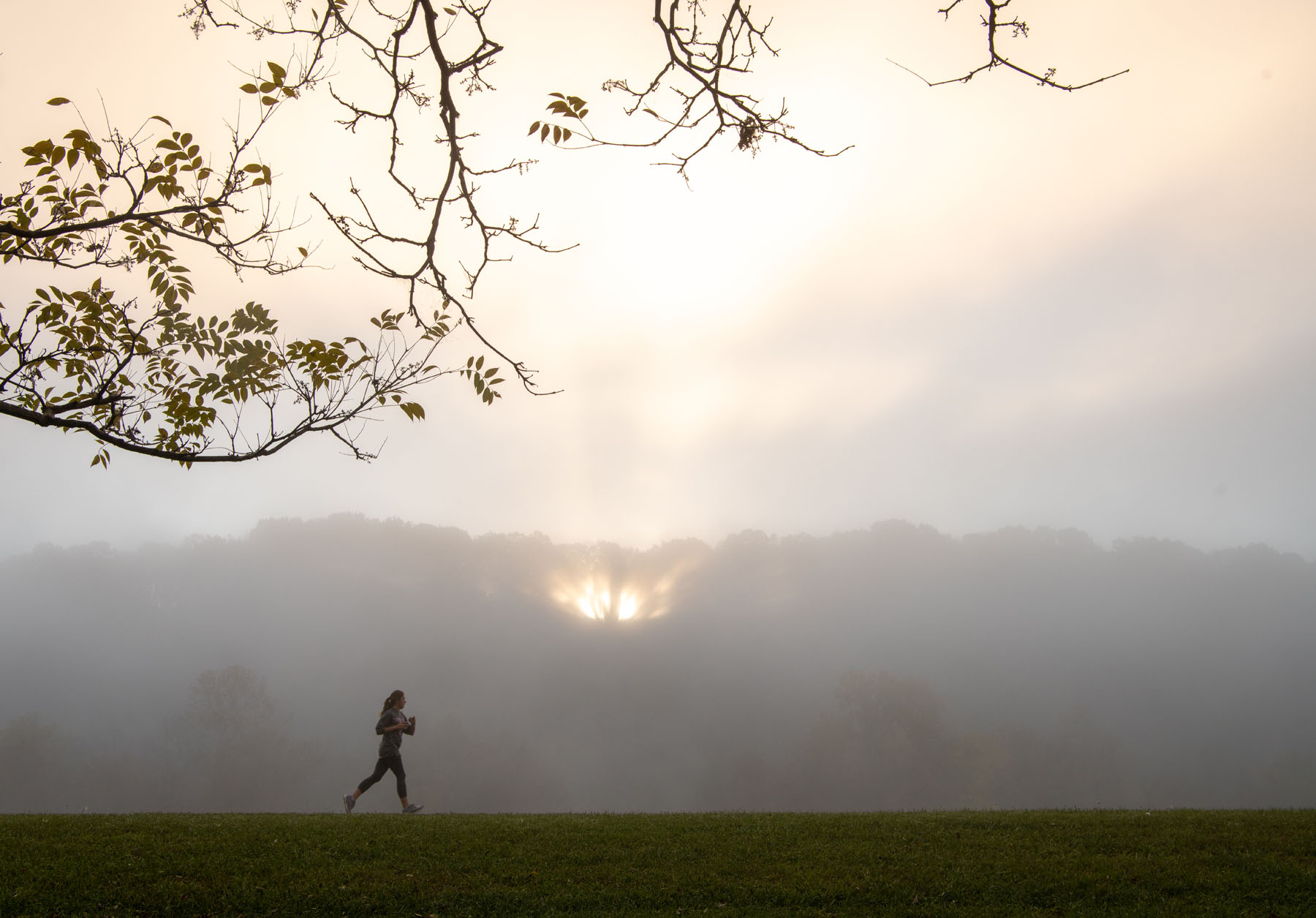 The sun rises over a foggy Hocking River as a jogger proceeds down the bike path parallel to the river