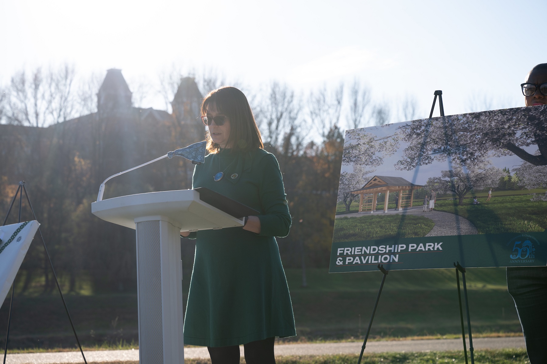 Gillian Ice is shown giving remarks near the sign for the Friendship Park and Pavilion